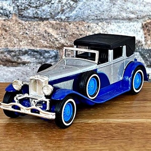 May include: A silver and blue vintage car model with a black fabric top. The car has a detailed interior and a chrome grill.