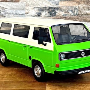 May include: A green and white toy Volkswagen van, a classic vehicle model. The van has a white roof and a bright green body. The front features a black grill with the Volkswagen logo. The van is on a wooden surface.