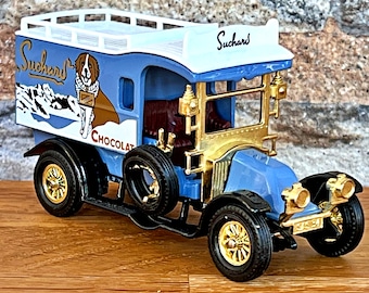 May include: A blue and white toy delivery truck with a gold trim. The truck is decorated with a mountain scene and a dog, and the words "Suchard Chocolat".