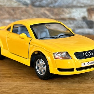Può includere: Un modello di auto giocattolo giallo di un'Audi TT Coupé. L'auto ha ruote nere e una griglia nera con il logo Audi.