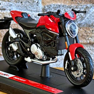 Peut inclure: Modèle réduit de moto Ducati Monster rouge et noir. Le modèle présente des pneus noirs, des tuyaux d'échappement argentés et le mot "Monster" sur le côté. Il est exposé sur une base noire avec le logo Ducati. Mesure environ 18 cm de long.
