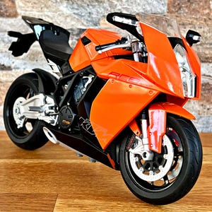 May include: Orange and black motorcycle model with a clear windshield. The motorcycle has a black seat and a silver exhaust pipe.