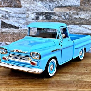 May include: A light blue vintage pickup truck model with white-walled tires and chrome accents. The toy truck is a classic design, with a detailed front grille and headlights. The truck is displayed on a wooden surface.