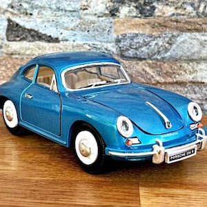 May include: A blue die-cast model of a Porsche 356 B coupe. The classic car has white-rimmed wheels, chrome bumpers, and a white stripe down the hood. The model is displayed on a wooden surface.