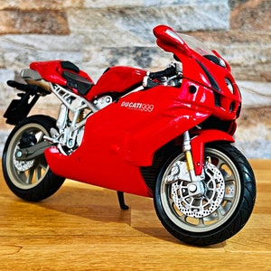 Peut inclure: Modèle de moto Ducati 999 rouge avec des accents noirs et des roues argentées. La moto est sur une surface en bois.