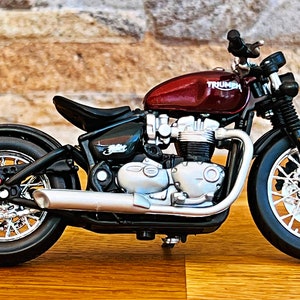 May include: A red Triumph motorcycle model with black wheels and a chrome exhaust pipe. The motorcycle is on a wooden surface.