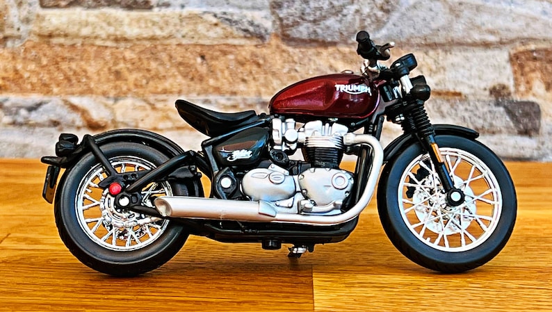 May include: A red Triumph motorcycle model with black wheels and a chrome exhaust pipe. The motorcycle is on a wooden surface.