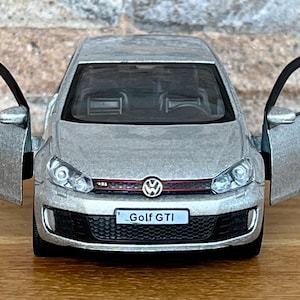 Puede incluir: Modelo plateado de un Volkswagen Golf GTI con las puertas abiertas. El coche tiene una parrilla negra, detalles rojos y el logotipo Golf GTI. Sobre un fondo de pared de ladrillo.