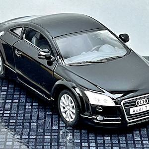 Puede incluir: Un coche modelo Audi TT negro con ruedas plateadas. El coche tiene un diseño elegante y aerodinámico con el logotipo de Audi y "Audi TT" en la parte delantera. El coche está sobre una superficie reflectante.