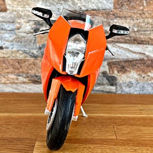 May include: Orange and black motorcycle model with a clear windshield and a black headlight. The motorcycle is on a wooden surface.