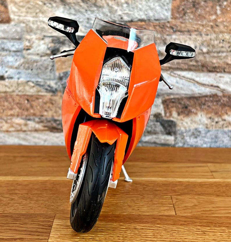 May include: Orange and black motorcycle model with a clear windshield and a black headlight. The motorcycle is on a wooden surface.