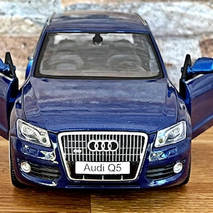 Könnte beinhalten: Ein blaues Audi Q5 Modellauto mit geöffneten Türen. Das Auto hat einen silbernen Kühlergrill und den Text "Audi Q5" auf der Vorderseite. Das Auto steht auf einer Holzoberfläche.