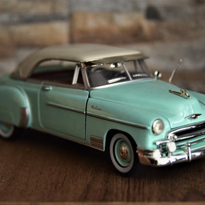 Könnte beinhalten: Ein blaugrünes und weißes Modellauto aus den 1950er Jahren mit Chrom-Akzenten. Das Auto hat ein weißes Dach und einen verchromten Kühlergrill. Das Auto ist ein Modell im Maßstab 1:18.