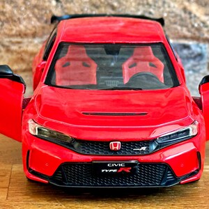 May include: A bright red die-cast model car featuring openable doors, a black front grille with a Honda emblem, and a number plate displaying the text CIVIC TYPE R. The interior includes red bucket seats and a black steering wheel.