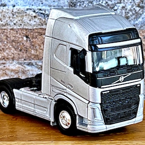 May include: A silver toy semi-truck with a black grill and a Volvo logo. The truck has a black cab and a silver trailer.