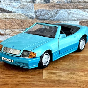 May include: A light blue Mercedes-Benz SL 500 convertible model car featuring a silver grille, clear headlights with orange turn signals, and a black interior. The front license plate displays the text S-SL 500 in black lettering on a white background.