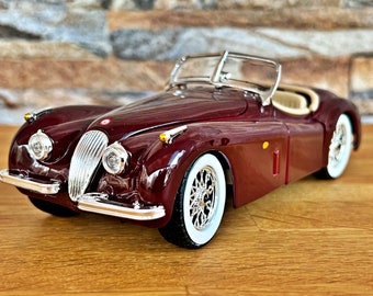 Op de afbeelding: Een bordeauxrode Jaguar XK120 roadster model auto uit de jaren 1950 met een wit cabriodak en chroomaccenten. De auto staat op een houten oppervlak.