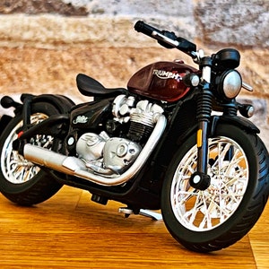 May include: A red and black Triumph motorcycle model with chrome accents. The motorcycle has a black seat and a black and chrome exhaust pipe.