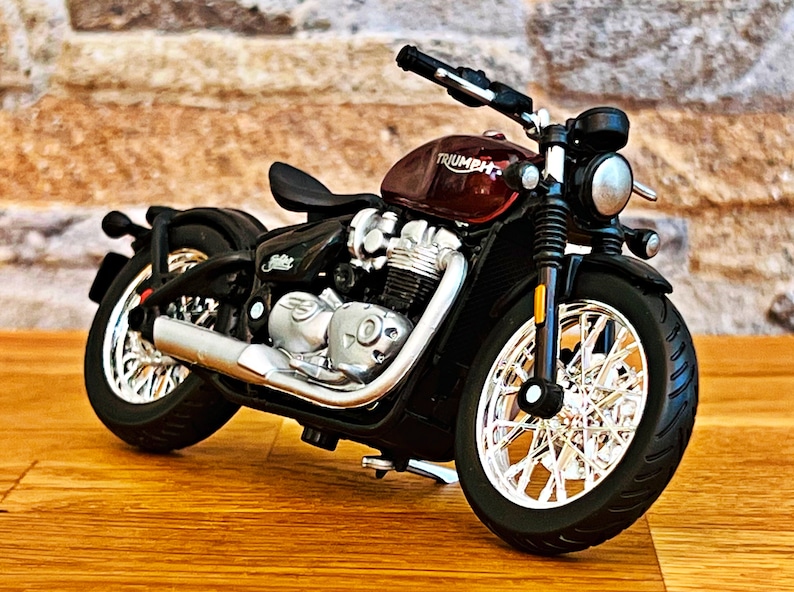 May include: A red and black Triumph motorcycle model with chrome accents. The motorcycle has a black seat and a black and chrome exhaust pipe.
