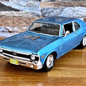 May include: A diecast model of a classic blue Chevrolet Nova SS. The car features chrome accents, a detailed interior, and a license plate. The model is displayed on a wooden surface.