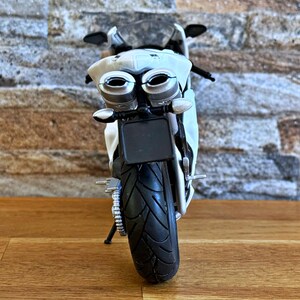 K&ouml;nnte beinhalten: Ein wei&szlig;es und silbernes Spielzeugmotorrad mit einem schwarzen Reifen und Nummernschild. Das Motorrad hat zwei Auspuffrohre und einen schwarzen Sitz. Der Hintergrund ist eine Backsteinmauer und eine Holzoberfl&auml;che.