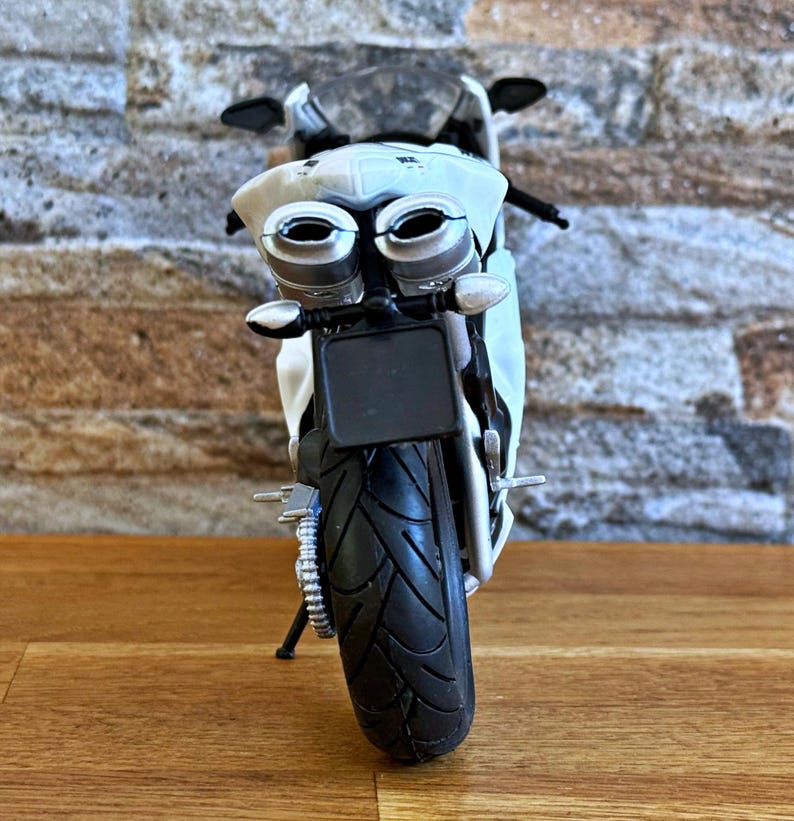 K&ouml;nnte beinhalten: Ein wei&szlig;es und silbernes Spielzeugmotorrad mit einem schwarzen Reifen und Nummernschild. Das Motorrad hat zwei Auspuffrohre und einen schwarzen Sitz. Der Hintergrund ist eine Backsteinmauer und eine Holzoberfl&auml;che.