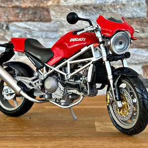 Peut inclure: Une moto Ducati rouge avec un cadre argenté et des roues noires. La moto a une selle noire et un pot d'échappement argenté. La moto est posée sur une surface en bois.