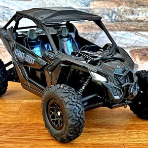 Puede incluir: Vehículo todoterreno Can-Am Maverick X3 negro de dos plazas con jaula antivuelco y ruedas negras.