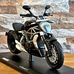 Peut inclure: Modèle réduit de moto Ducati X Diavel S noire. Le modèle présente un cadre, des roues et une carrosserie noirs avec des détails argentés. La moto est exposée sur un socle noir avec le logo Ducati et le texte "X Diavel S".