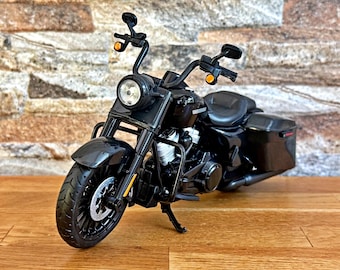 May include: A black scale model motorcycle featuring a prominent round headlight, high handlebars with side mirrors, orange turn signals, a detailed engine block, a single seat, and hard-shell saddlebags, supported by a kickstand on a wooden surface.