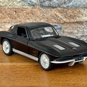 May include: A black die-cast model of a classic sports car with silver trim and wheels. The car has a sleek, aerodynamic design with a low profile and a hardtop. The model is displayed on a wooden surface.