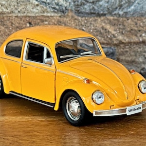 May include: A bright yellow VW Beetle model car. The classic car has black tires, chrome accents, and the words "VW Beetle" on the front. The car is sitting on a wooden surface.