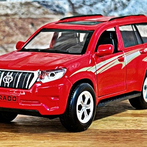 May include: Red toy car model of a Toyota Land Cruiser Prado SUV. The car has a black roof rack, silver accents, and a black grille with the word "PRADO" printed on it.