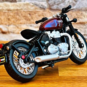May include: A red and silver toy motorcycle with the word "Triumph" on the side. The motorcycle is on a wooden surface.