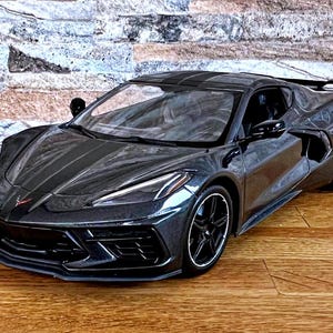 May include: A dark gray sports car with black wheels and a black stripe down the center of the hood. The car is parked on a wooden floor.