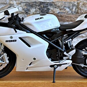 K&ouml;nnte beinhalten: Wei&szlig;es Ducati 1198 Motorradmodell mit schwarzen Akzenten. Das Modell verf&uuml;gt &uuml;ber einen detaillierten Motor, R&auml;der und Auspuffrohre. Das Motorrad wird auf einer Holzoberfl&auml;che vor einer Backsteinmauer gezeigt.