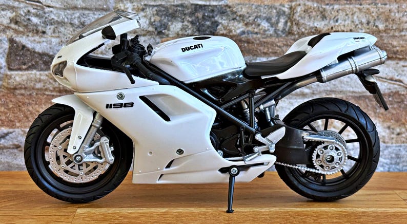 K&ouml;nnte beinhalten: Wei&szlig;es Ducati 1198 Motorradmodell mit schwarzen Akzenten. Das Modell verf&uuml;gt &uuml;ber einen detaillierten Motor, R&auml;der und Auspuffrohre. Das Motorrad wird auf einer Holzoberfl&auml;che vor einer Backsteinmauer gezeigt.