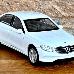 May include: A white sedan model car with silver rims and black tires. The car has a black interior and tinted windows. The front of the car features a chrome emblem. The car is sitting on a wooden surface.
