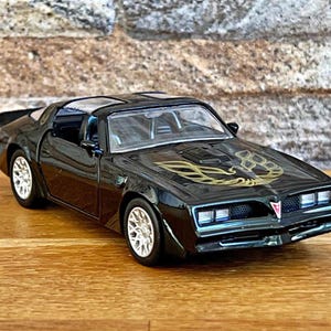May include: A black and gold die-cast model car of a Pontiac Trans Am. The car has a black body with gold accents and a gold bird logo on the hood.