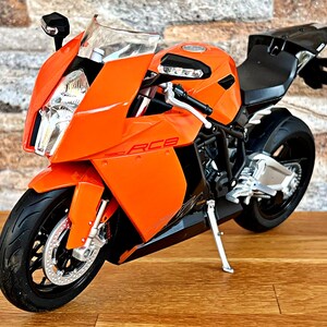 May include: Orange and black motorcycle model with a white and silver front wheel. The motorcycle has a black seat and a silver exhaust pipe.