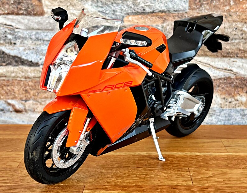May include: Orange and black motorcycle model with a white and silver front wheel. The motorcycle has a black seat and a silver exhaust pipe.