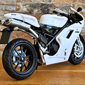 K&ouml;nnte beinhalten: Wei&szlig;es Ducati 1198 Motorradmodell mit schwarzen R&auml;dern und Akzenten. Das Modell verf&uuml;gt &uuml;ber ein detailliertes Design, einschlie&szlig;lich Auspuffrohren, einer Windschutzscheibe und dem Ducati-Logo. Das Motorrad steht auf einer Holzoberfl&auml;che.
