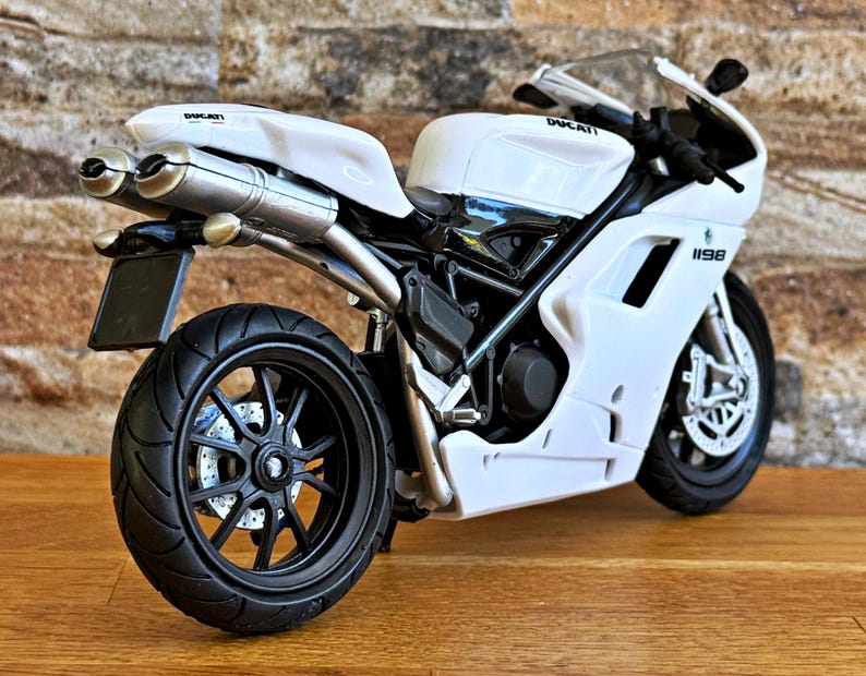 K&ouml;nnte beinhalten: Wei&szlig;es Ducati 1198 Motorradmodell mit schwarzen R&auml;dern und Akzenten. Das Modell verf&uuml;gt &uuml;ber ein detailliertes Design, einschlie&szlig;lich Auspuffrohren, einer Windschutzscheibe und dem Ducati-Logo. Das Motorrad steht auf einer Holzoberfl&auml;che.