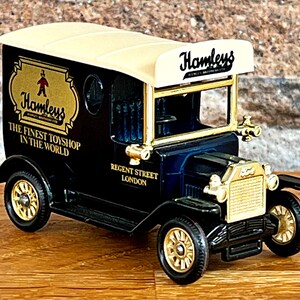 May include: A black toy delivery van with gold accents and the words "Hamleys The Finest Toyshop in the World" on the side. The van has a white roof with the word "Hamleys" in gold lettering. The van is on a wooden surface.