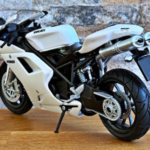 K&ouml;nnte beinhalten: Ein wei&szlig;es Ducati 1198 Motorradmodell mit schwarzen Akzenten. Das Modell hat ein schlankes Design, sichtbare Auspuffrohre und das Ducati-Logo. Das Motorrad steht auf einer Holzoberfl&auml;che vor einer Backsteinmauer.