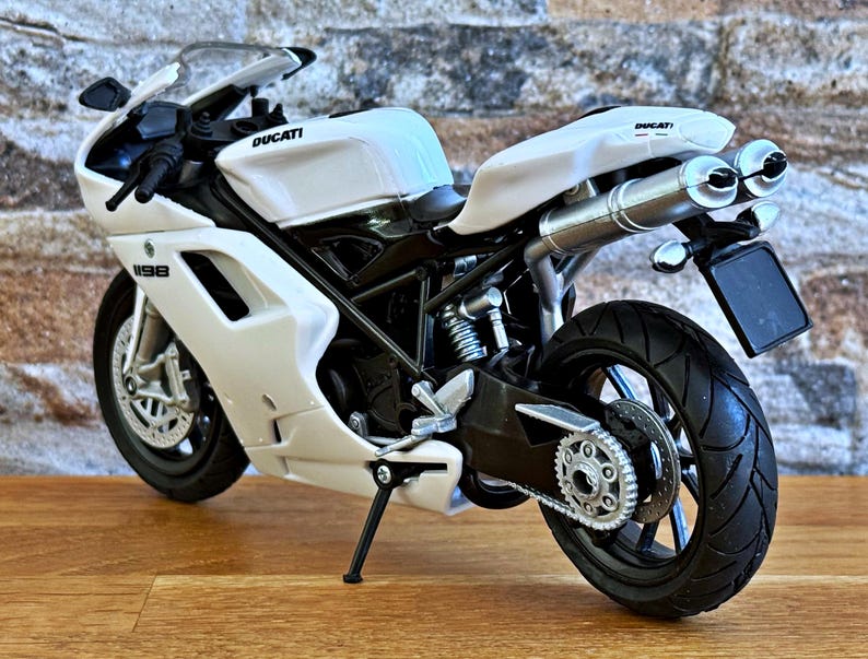 K&ouml;nnte beinhalten: Ein wei&szlig;es Ducati 1198 Motorradmodell mit schwarzen Akzenten. Das Modell hat ein schlankes Design, sichtbare Auspuffrohre und das Ducati-Logo. Das Motorrad steht auf einer Holzoberfl&auml;che vor einer Backsteinmauer.