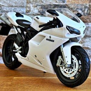 K&ouml;nnte beinhalten: Ein wei&szlig;es Ducati 1198 Motorradmodell mit schwarzen Akzenten. Das Modell hat ein schlankes Design, schwarze Reifen und eine klare Windschutzscheibe. Das Motorrad steht vor einer strukturierten Backsteinmauer und einer Holzoberfl&auml;che.