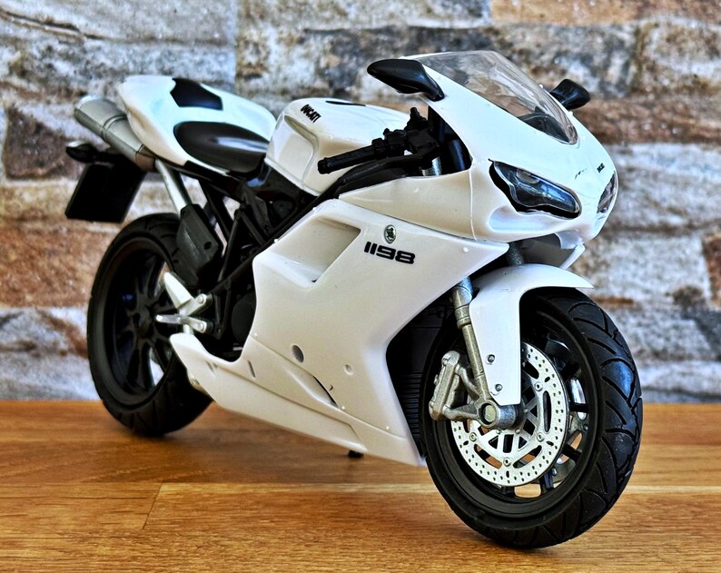 K&ouml;nnte beinhalten: Ein wei&szlig;es Ducati 1198 Motorradmodell mit schwarzen Akzenten. Das Modell hat ein schlankes Design, schwarze Reifen und eine klare Windschutzscheibe. Das Motorrad steht vor einer strukturierten Backsteinmauer und einer Holzoberfl&auml;che.