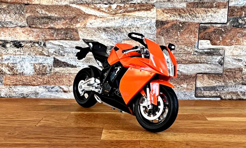 May include: Orange and black sport motorcycle with a black seat and a black windshield. The motorcycle is on a wooden floor in front of a brick wall.