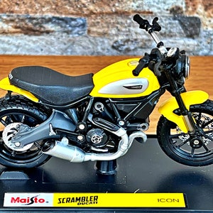 Peut inclure: Modèle de moto Ducati Scrambler jaune avec des roues noires et une selle noire. La moto est sur un présentoir noir avec une base jaune sur laquelle est inscrit "Maisto" et "Scrambler Ducati" en noir. La base indique également "ICON" en noir.